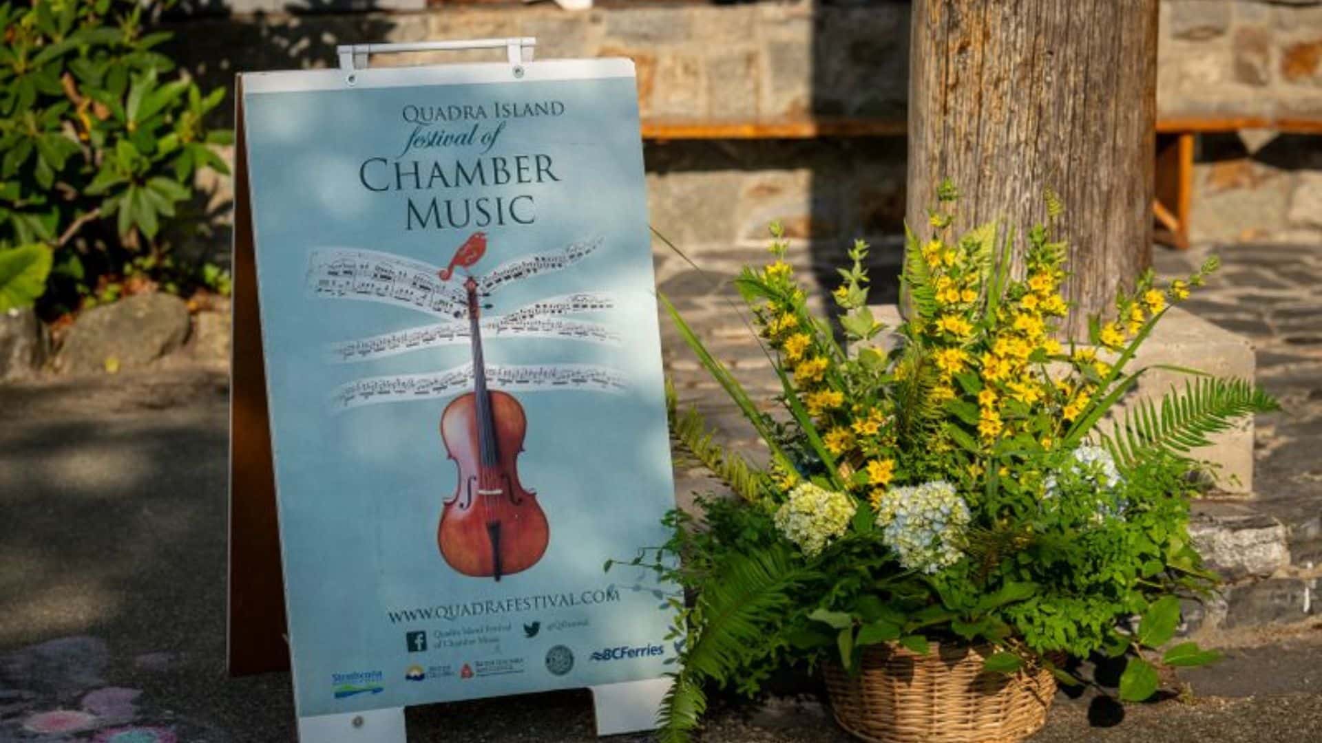 A poster for the Quadra Island Festival of Chamber Music stands outdoors beside yellow and white flowers. The poster features a violin with musical notes, a bird perched on top, and details on how to get your Festival Pass.