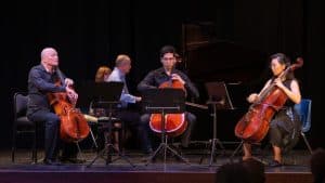 Three cellists sit on stage playing their instruments in an atmosphere of Intimate Conversations, with sheet music stands before them. In the background, two pianists share the grand piano against a dark backdrop.