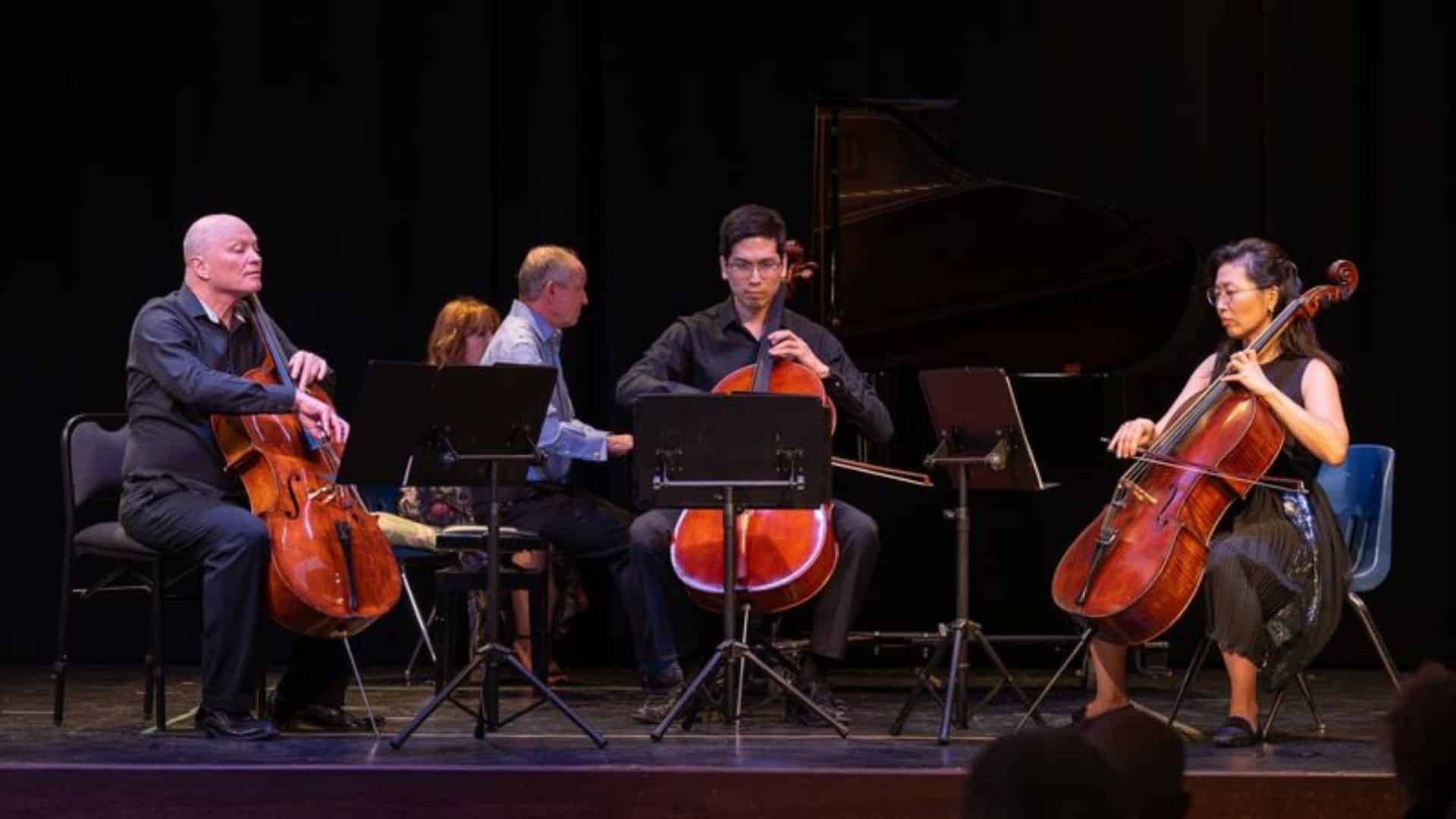 Three cellists sit on stage playing their instruments in an atmosphere of Intimate Conversations, with sheet music stands before them. In the background, two pianists share the grand piano against a dark backdrop.