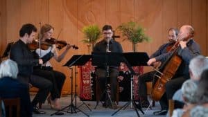 Five musicians perform indoors, playing violin, viola, cello, and clarinet, seated in a semicircle. Audience members watch in the foreground. The background features wood paneling and decorative plants.