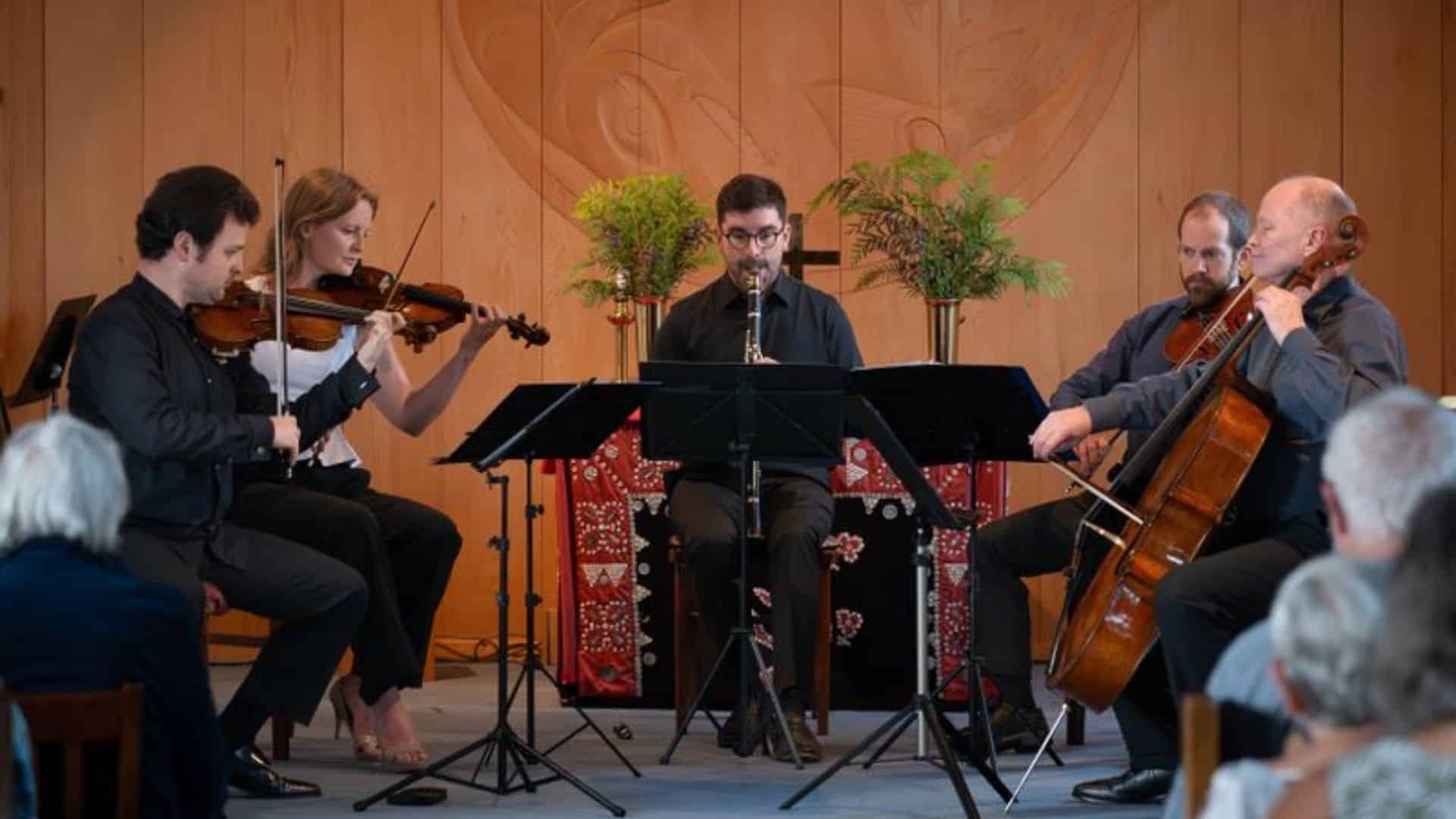 Five musicians perform indoors, playing violin, viola, cello, and clarinet, seated in a semicircle. Audience members watch in the foreground. The background features wood paneling and decorative plants.