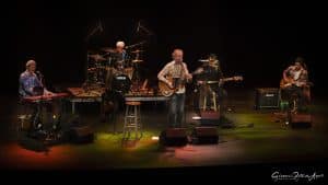 A five-member band performs on a dimly lit stage for a Gordon Lightfoot tribute show, featuring a keyboardist, drummer, two guitarists, and a bassist, surrounded by microphones, amps, and music stands.