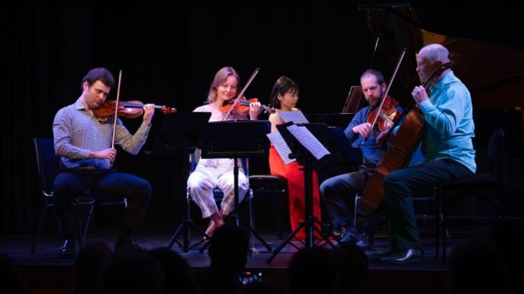 Five musicians perform on stage with string instruments and a piano, reading from sheet music stands. Seated in a semicircle under stage lighting, spark and shadow shimmer across the dark background as the audience watches.