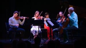 Five musicians perform on stage with string instruments and a piano, reading from sheet music stands. Seated in a semicircle under stage lighting, spark and shadow shimmer across the dark background as the audience watches.