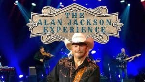 A man in a cowboy hat and embroidered shirt stands before a band playing on stage, with a large sign above proudly announcing The Alan Jackson Experience in decorative lettering.