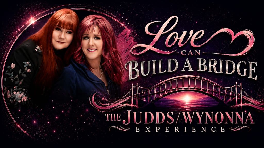 Two women with red hair smile beside the text "Love Can Build A Bridge – The Judds/Wynonna Experience," featuring a pink bridge and sunset design on a dark, starry background.