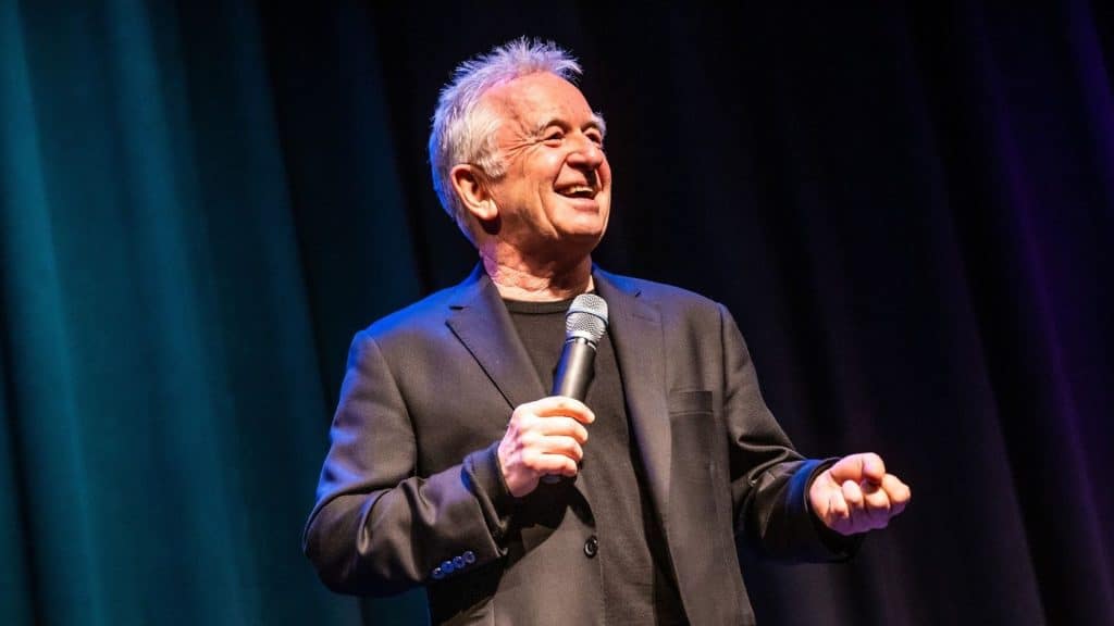 An older man with gray hair, wearing a black suit and t-shirt, smiles and holds a microphone while performing comedy onstage in front of dark curtains with blue and purple lighting.