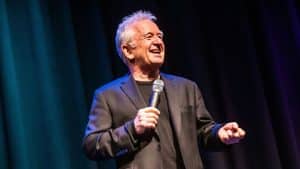 An older man with gray hair, wearing a black suit and t-shirt, smiles and holds a microphone while performing comedy onstage in front of dark curtains with blue and purple lighting.
