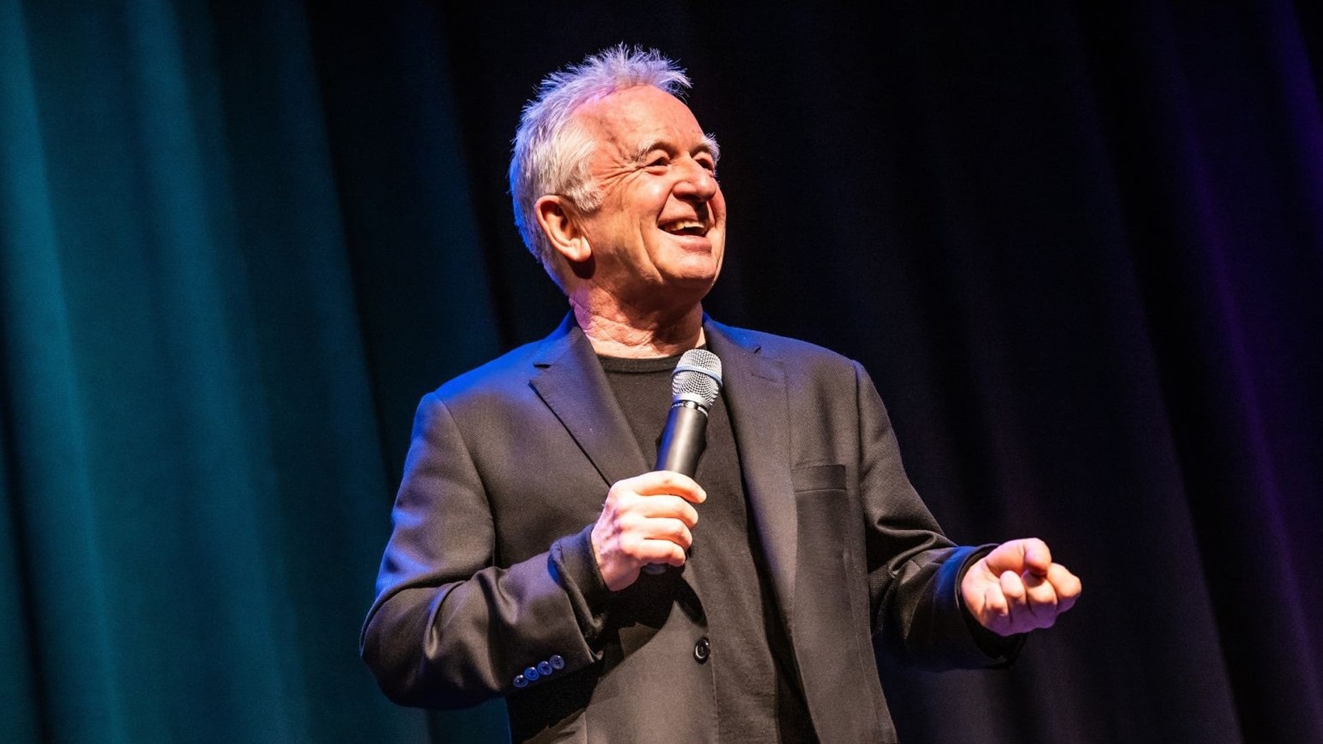 An older man with gray hair, wearing a black suit and t-shirt, smiles and holds a microphone while performing comedy onstage in front of dark curtains with blue and purple lighting.