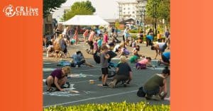 People of all ages draw colorful chalk art on a closed city street during the River City Streets Fest. Trees, buildings, and a white tent line the background. The CR Live Streets logo appears in the top left corner.