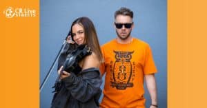A woman playing a black electric violin with a bow smiles at the camera while a man in sunglasses and an orange t-shirt stands behind her against a gray wall. The image has an orange border with the "CR Live Streets" logo in the top left corner.
