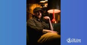 A bearded man in a dark shirt and cap sits on a couch under a lamp, smiling and relaxed. The softly lit background adds to the Country vibe. The CR Live Streets logo appears in the bottom right corner on a blue border.