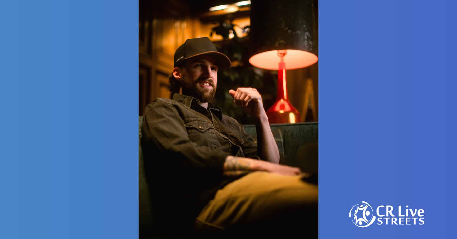 A bearded man in a dark shirt and cap sits on a couch under a lamp, smiling and relaxed. The softly lit background adds to the Country vibe. The CR Live Streets logo appears in the bottom right corner on a blue border.
