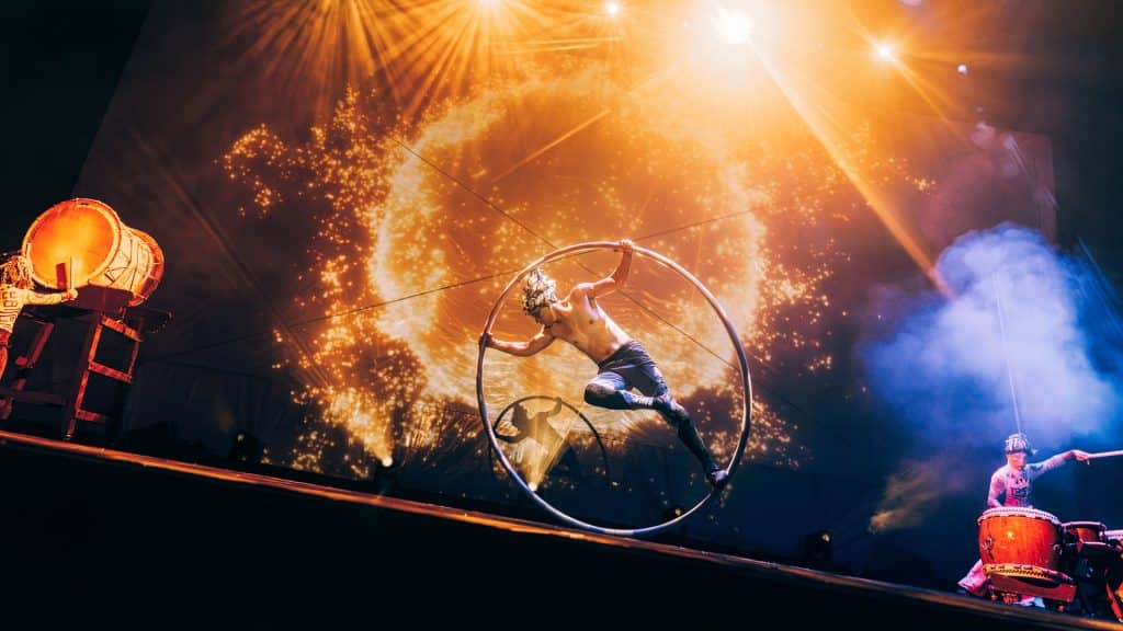 A shirtless performer from Diabolo Dance Theatre spins inside a large metal hoop on stage, with dramatic lighting and a fiery circular visual in the background. Two drummers play on either side, infusing the energetic scene with the spirit of Taiwan.