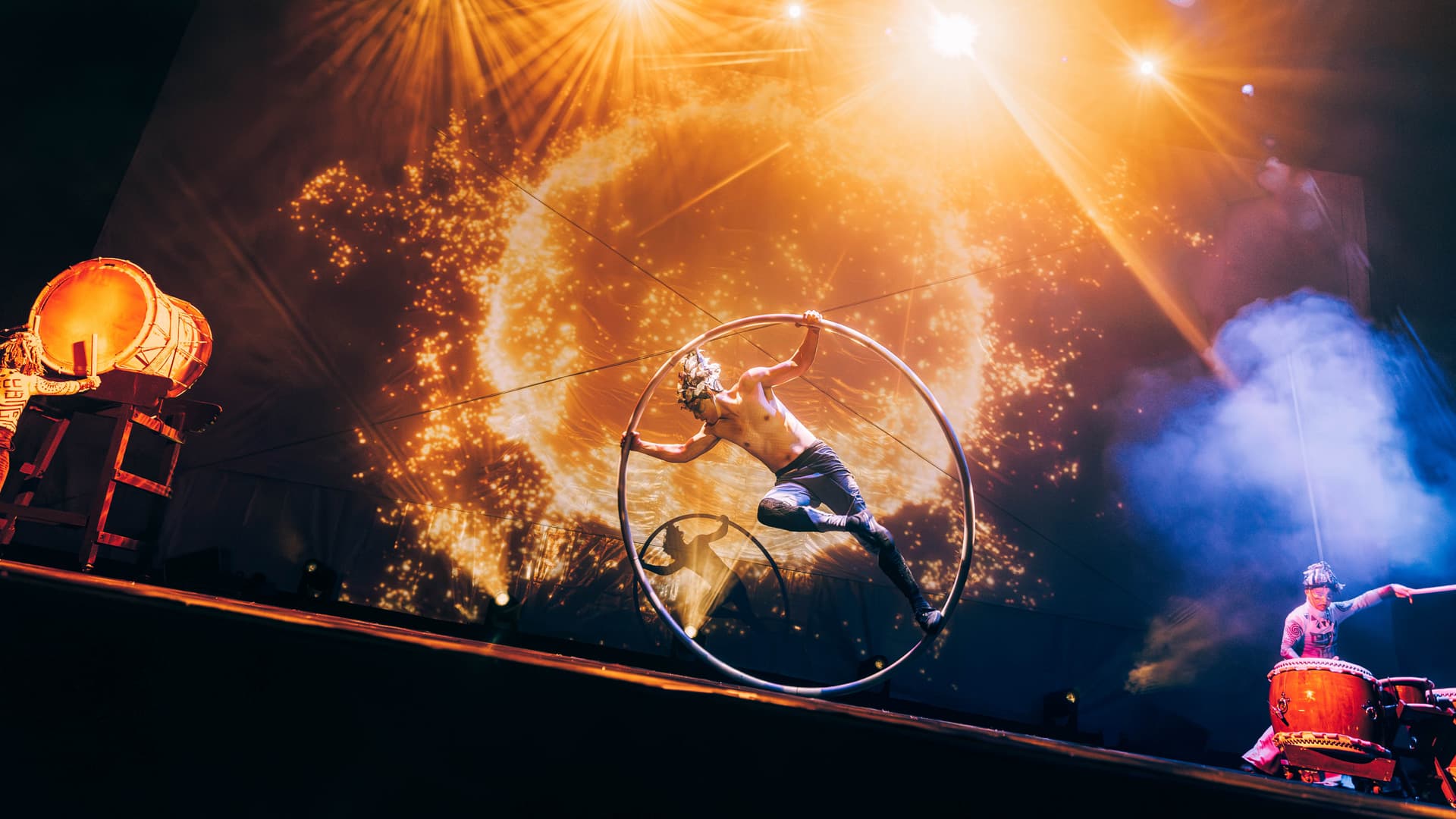 A shirtless performer from Diabolo Dance Theatre spins inside a large metal hoop on stage, with dramatic lighting and a fiery circular visual in the background. Two drummers play on either side, infusing the energetic scene with the spirit of Taiwan.