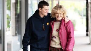 A man in a navy hoodie smiles with his arm around a woman in glasses and a pink jacket; both are walking outside and appear happy, sharing an “I Swear” moment that captures genuine joy.