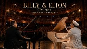 Two musicians play black and white grand pianos on a theater stage before a large audience, with the text “BILLY & ELTON: The Legacy. Two Pianos. One Night.” displayed above them, celebrating the timeless music of Billy & Elton.