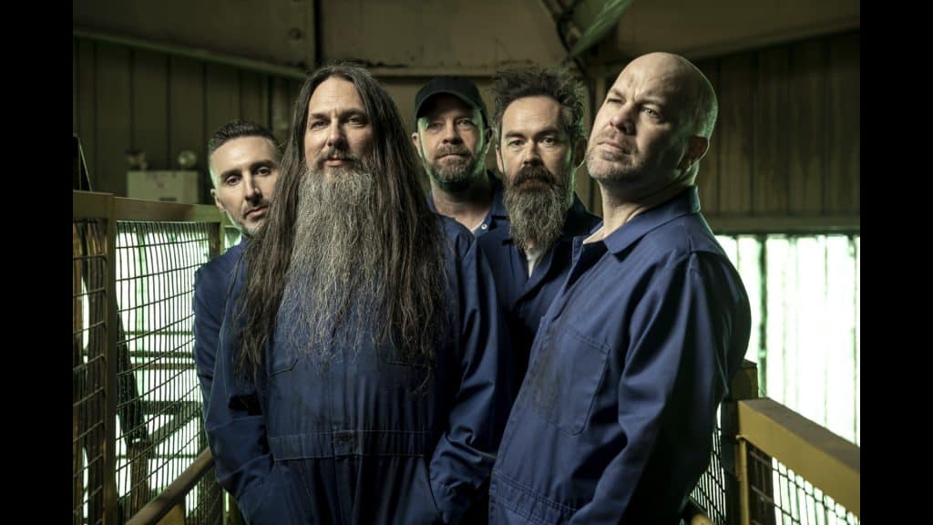 Five men in blue coveralls stand close together in an industrial setting with metal railings, two with long hair and beards, all looking towards the camera with serious expressions—much like the members of Finger Eleven preparing for a powerful performance.