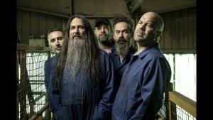 Five men in blue coveralls stand close together in an industrial setting with metal railings, two with long hair and beards, all looking towards the camera with serious expressions—much like the members of Finger Eleven preparing for a powerful performance.