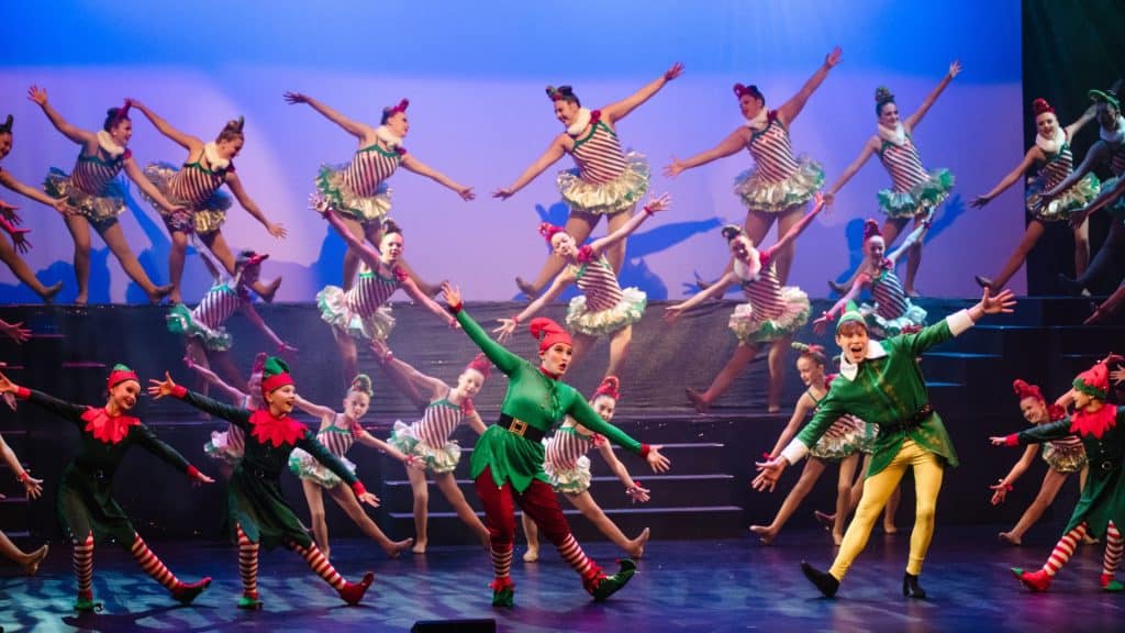 A troupe of performers in elf costumes dancing on stage during the A Clause To Remember Christmas Spectacular, with two lead elves positioned in the foreground.