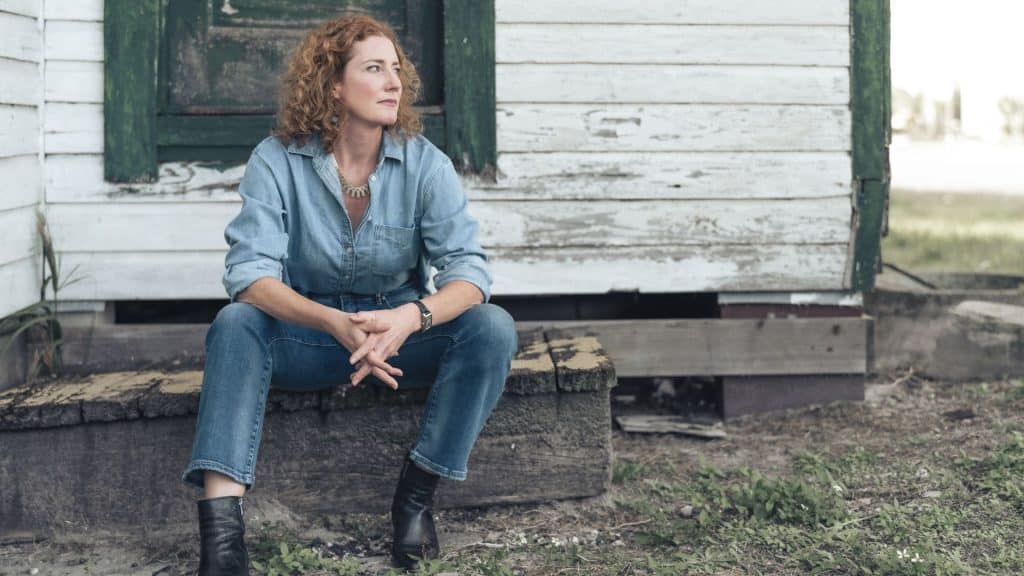 Musician Kathleen Edwards with curly red hair sits on the steps of a weathered wooden building.