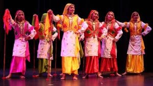 Six dancers in vibrant traditional outfits embody the spirit of India as they perform on a stage, holding props and wearing headscarves.